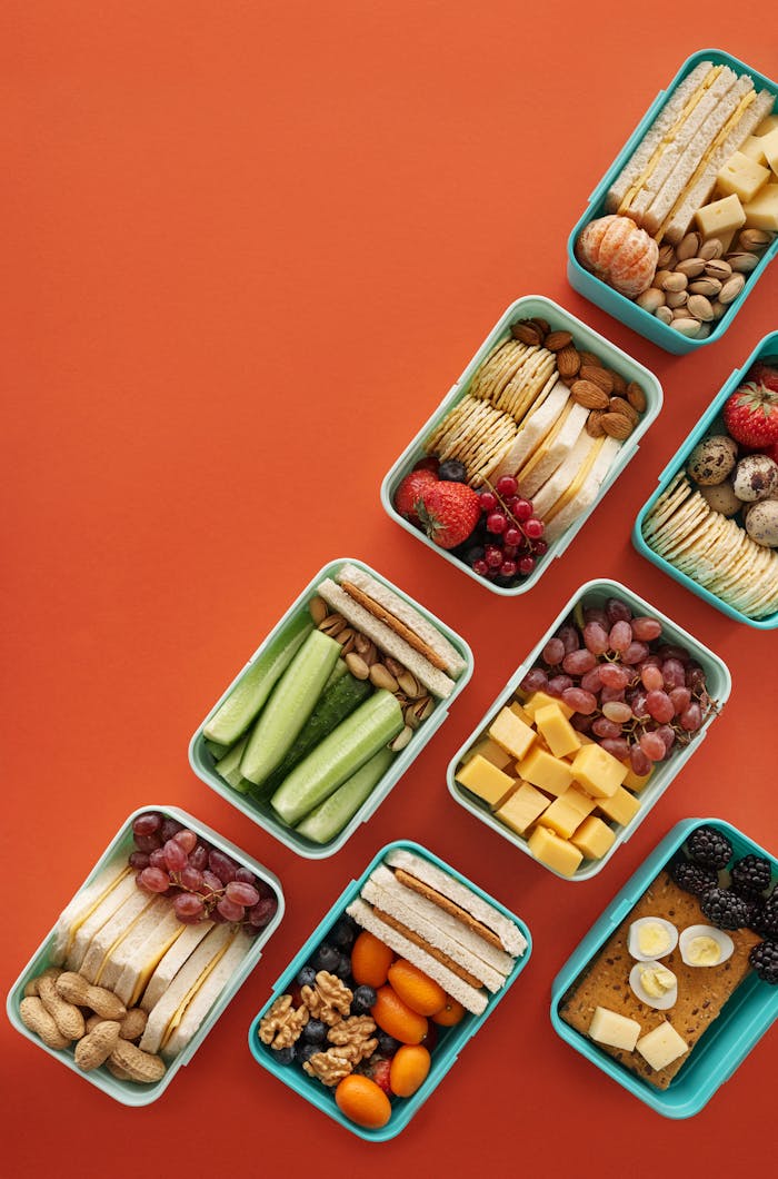services-01 Flat lay of assorted healthy lunchboxes with fruits and nuts against vibrant orange background.