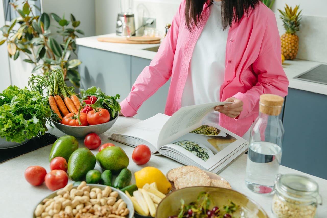 journey A person browsing a cookbook surrounded by fresh vegetables in a bright kitchen setting.